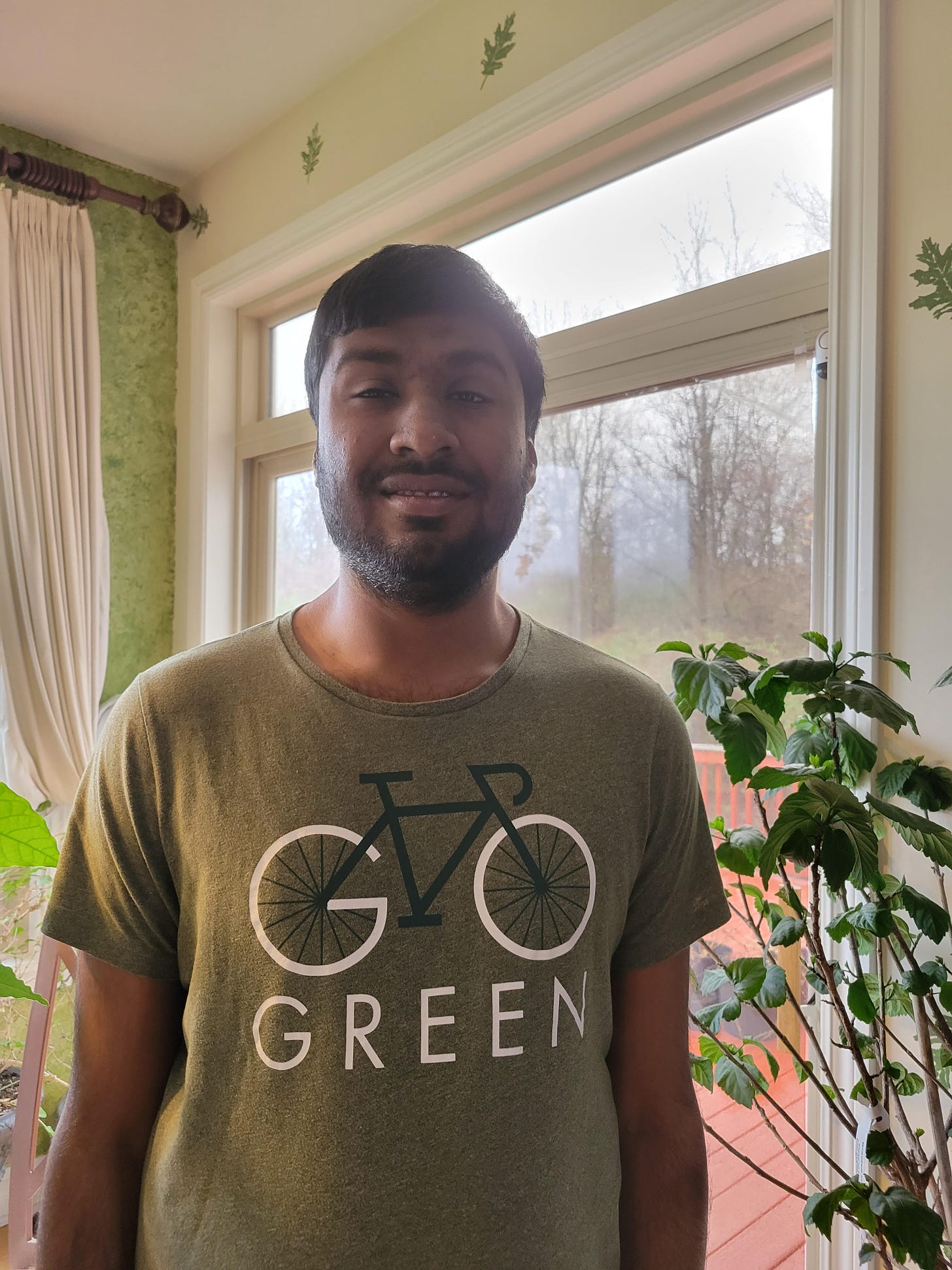Kashi Krishnan appears before a window in a close-cut beard and a green shirt that says go green. 