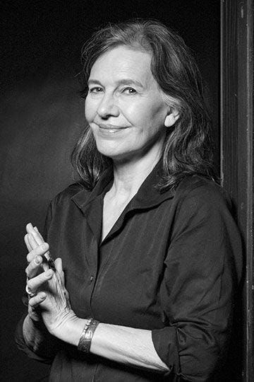 Black & white portrait of Louise Erdrich from the waist up, taken at a slight angle. She is smiling like she has a secret.