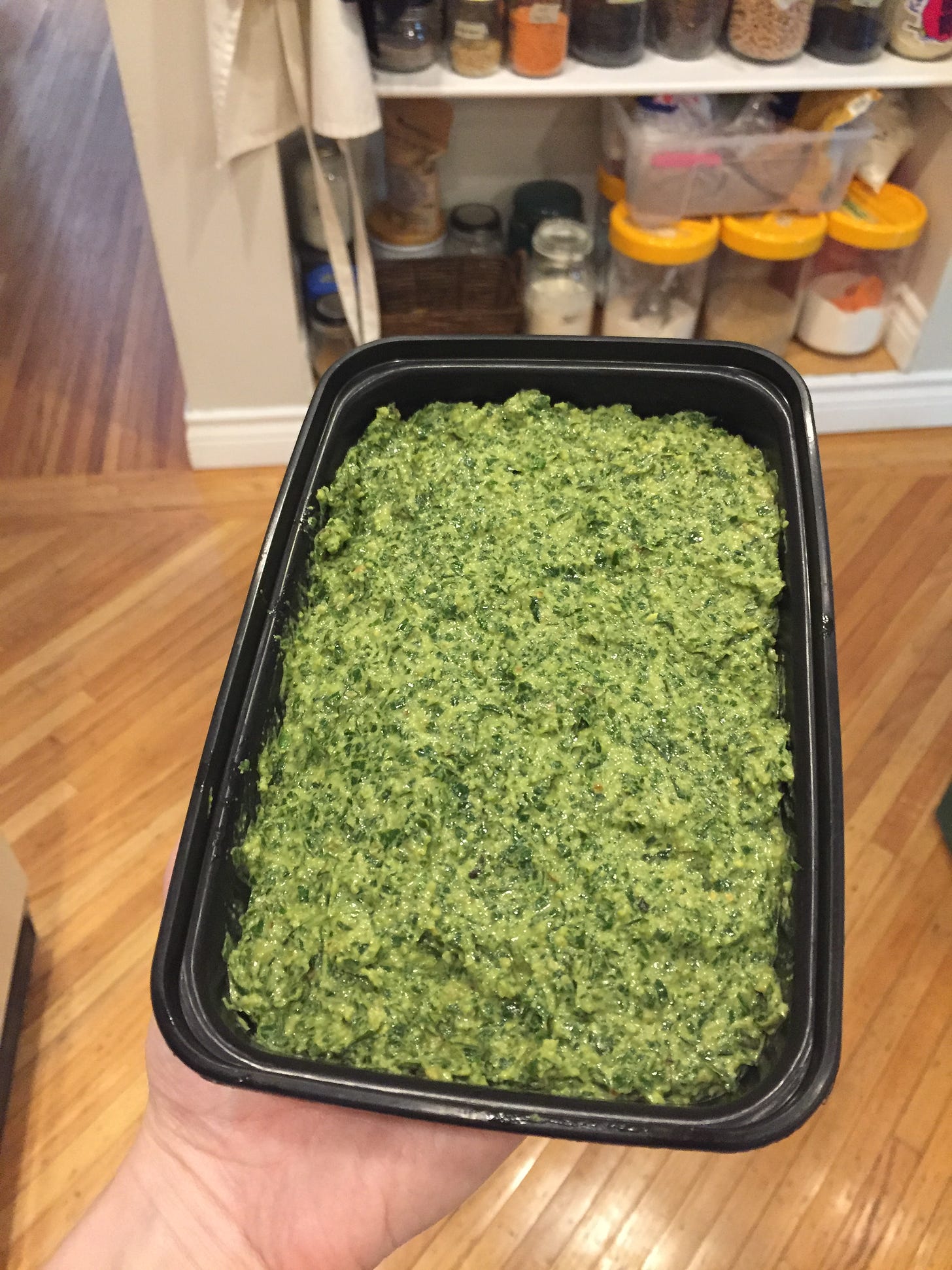 a black rectangular container full of pesto, held over a wood floor.