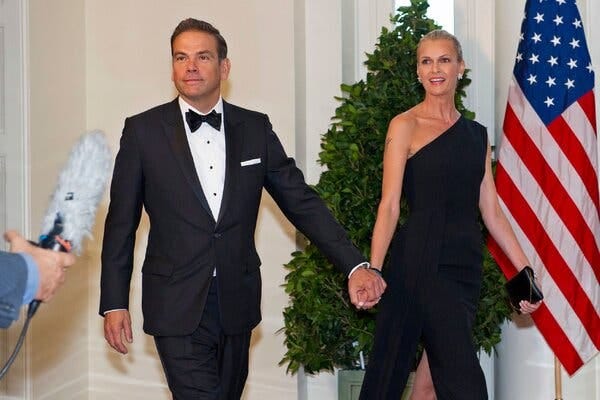 Lachlan Murdoch, wearing a black tuxedo and bow tie, holds hands with his wife, wearing a long black dress.
