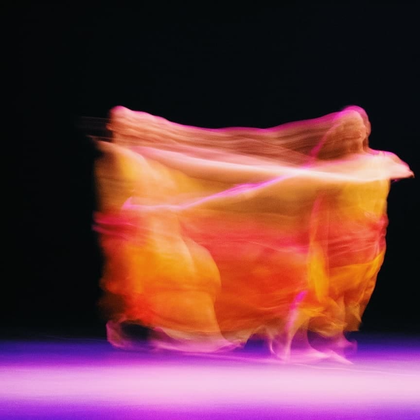 Fotografia em longa exposição de uma dançarina. Ela está em palco, o fundo é escuro. Ela veste uma roupa amarela, rosa e roxa. A imagem forma um borrão dessas cores.