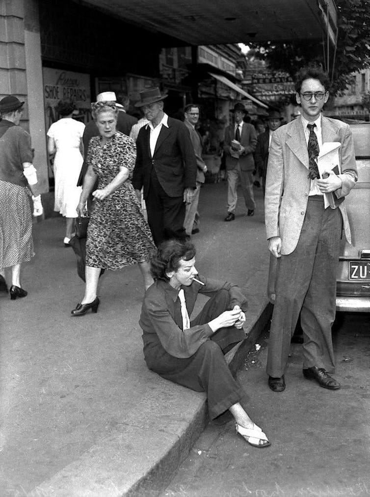 Norton wearing slacks, smoking in public, sitting in the gutter. A woman walks past, looking scandalised.