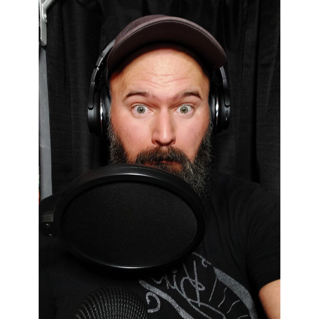 A shoulders-up selfie of a white man with a thick salt and pepper beard, green eyes, and grey ball cap makes a faux scared face. His chin is partially obscured by a microphone and he is wearing headphones. He's inside of a small audio studio with black curtains for wall.