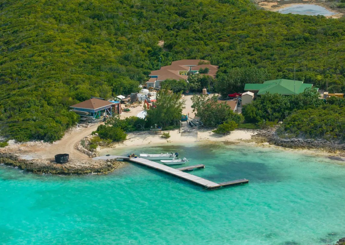 A complex of several large buildings set very close to a private beach on a tropical island, surrounded by trees and greenery. The water by the beach is a transparent aquamarine. There is a small wooden pier with two little boats moored off them.