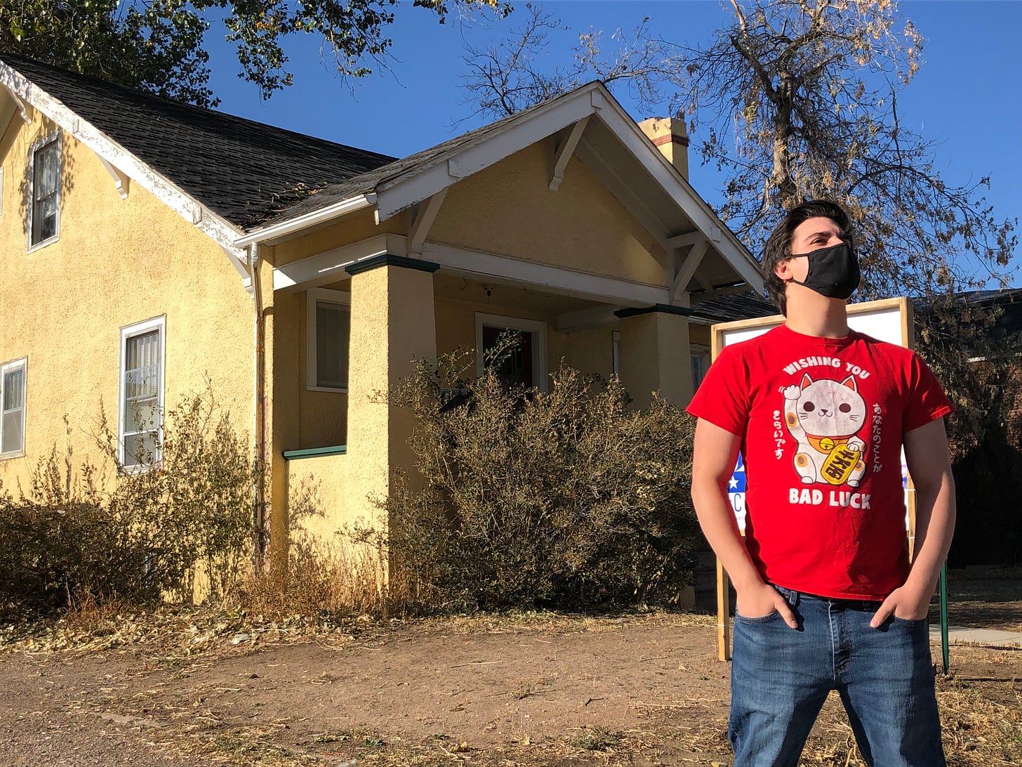 A young man in a red t-shirt and black mask stands in front of a yellow house.