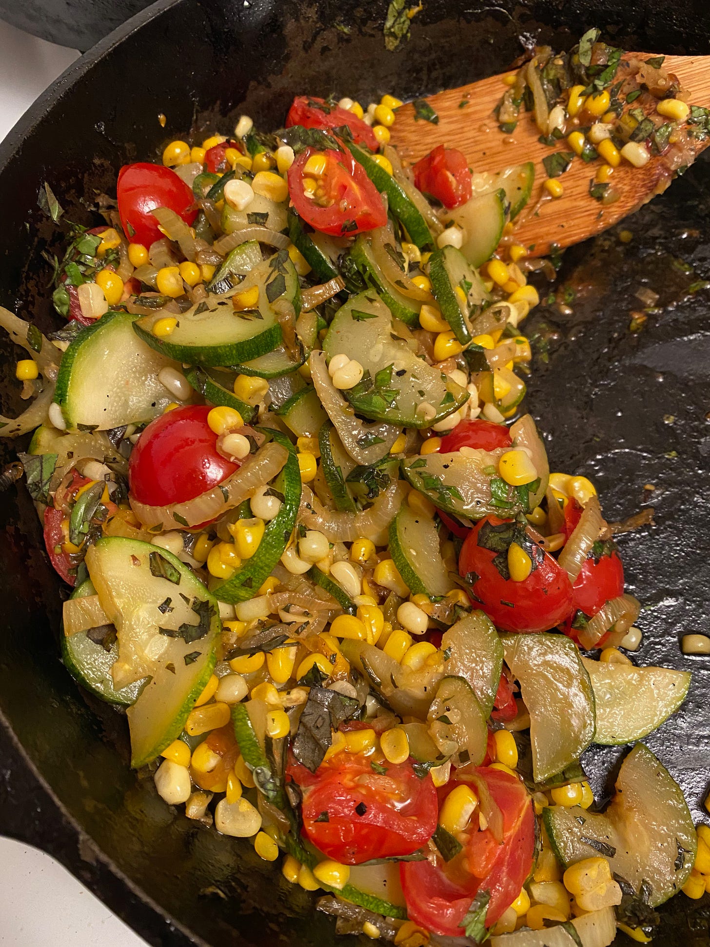 A veggie sauté in a cast-iron frying pan. The veggies — zucchini, corn, tomatoes, onions and fennel — are all scooped to one side of the pan, and a wooden spatula is visible in the top corner.
