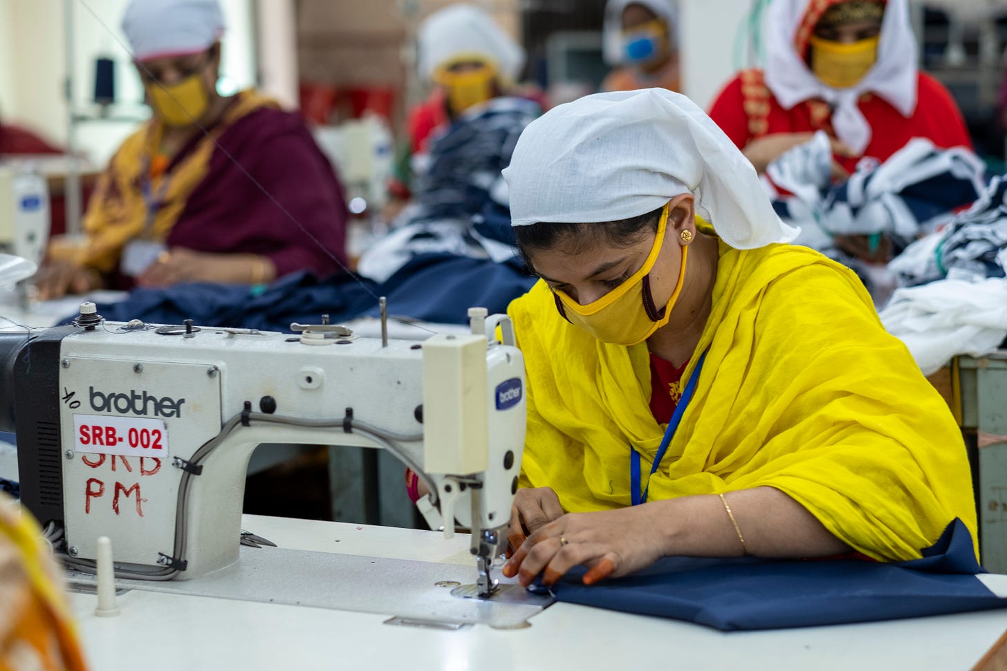 A woman in a yellow sari and facemask works at a sewing machine in a factory, surrounded by other women doing the same