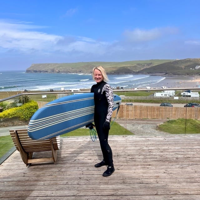 A woman in a wetsuit, stading on a wooden deck holding a surfboard and smiling, a beach and ocean in the background