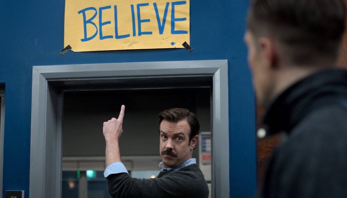 Ted Lasso pointing at a sign above his office door that reads "Believe".