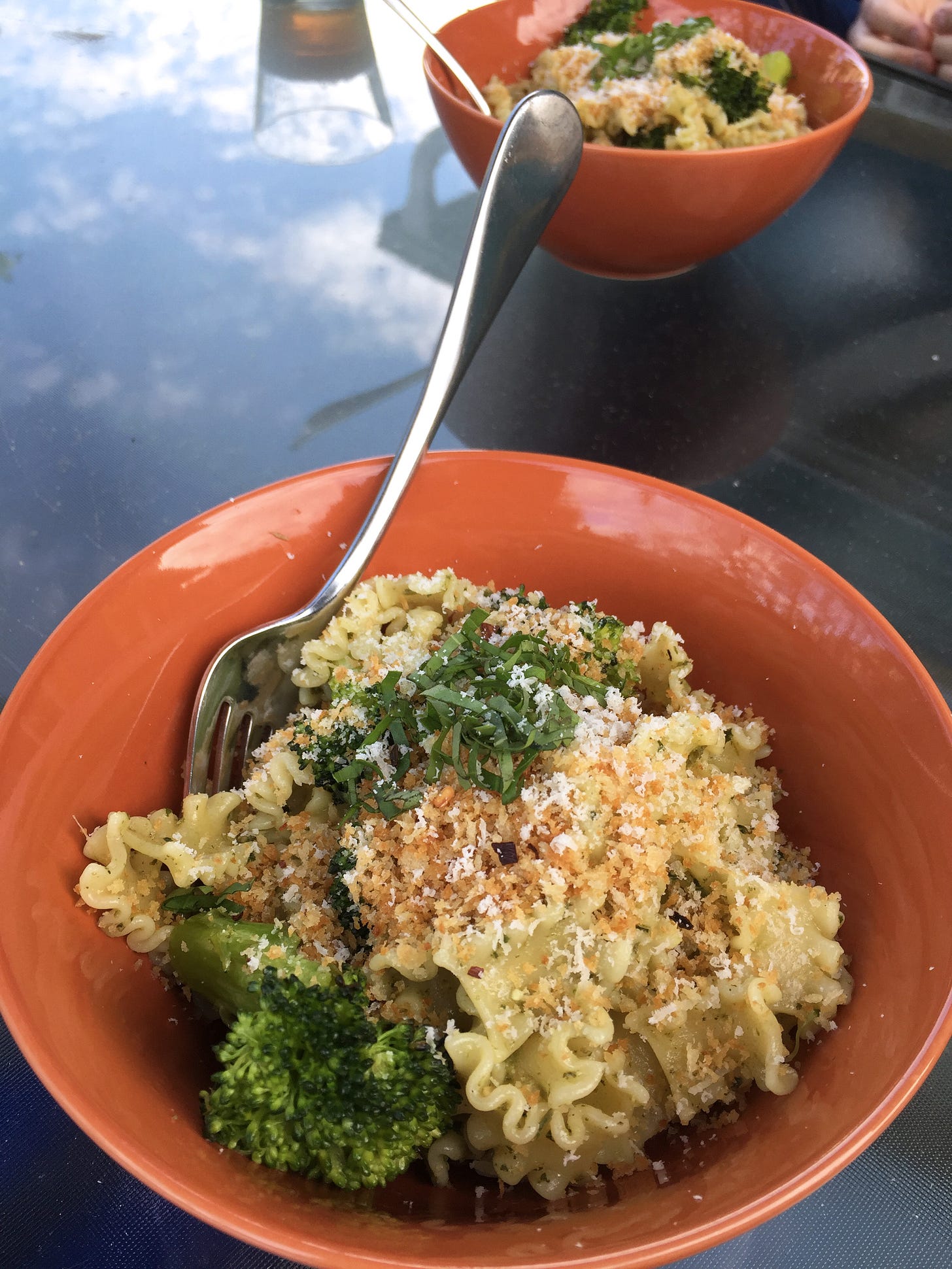 two orange bowls filled with pesto-coated mafalda noodles, with pieces of charred broccoli. On top are toasted panko crumbs with shreds of parmesan and basil.