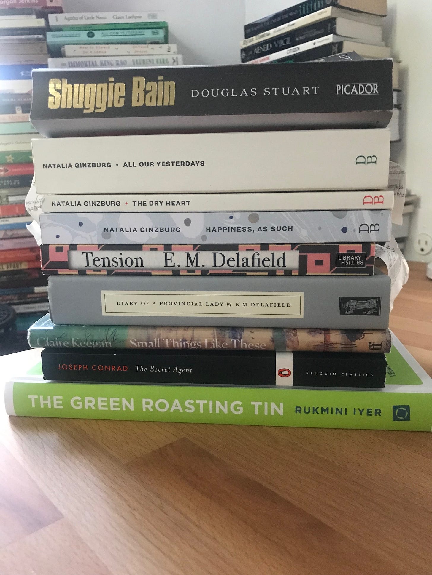 Stack of books purchased on vacation. Includes Shuggie Bain, 3 by Ginzburg, 2 by Delafield, a vegetarian cookbook, "Small Things Like These," and "The Secret Agent."