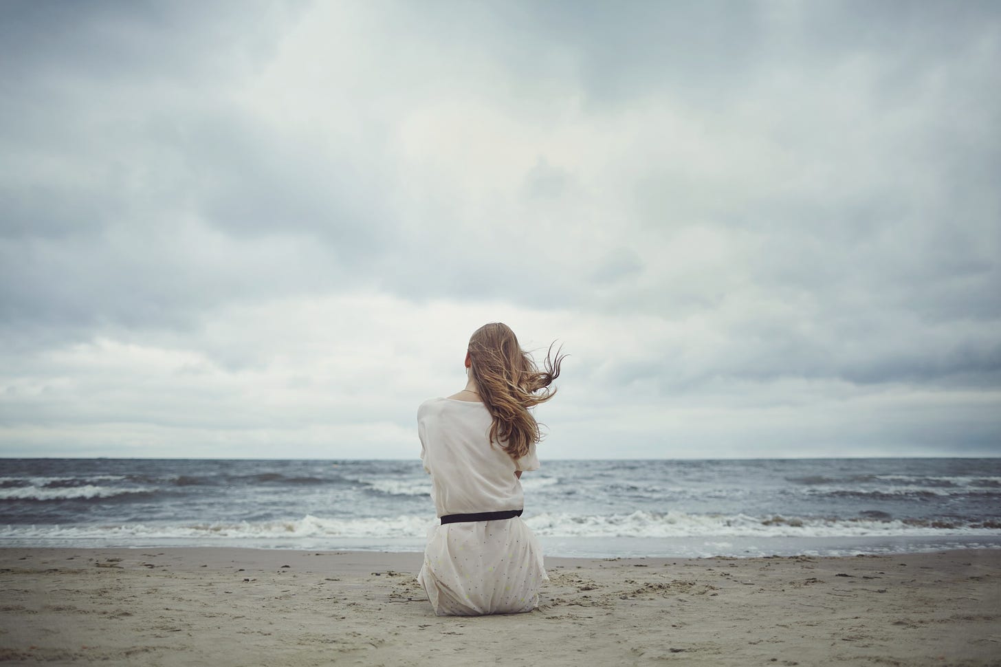 A woman sits alone on a windy beach as a storm comes in.