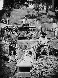 iew of 4 men, two miners at work on a long tom (one man is an African American), one man in a wagon, one man standing behind the wagon.