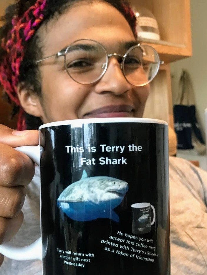 Picture of Chigozie Nri smiling and holding up a coffee mug that says “This is Terry the Fat Shark” above a picture of a shark that does indeed look portly. Below Terry it says “He hopes you will accept this coffee mug printed with Terry’s likeness as a token of friendship” and “Terry will return with another gift next Wednesday.” And incredibly, in front of Terry there is a picture of this very mug. 