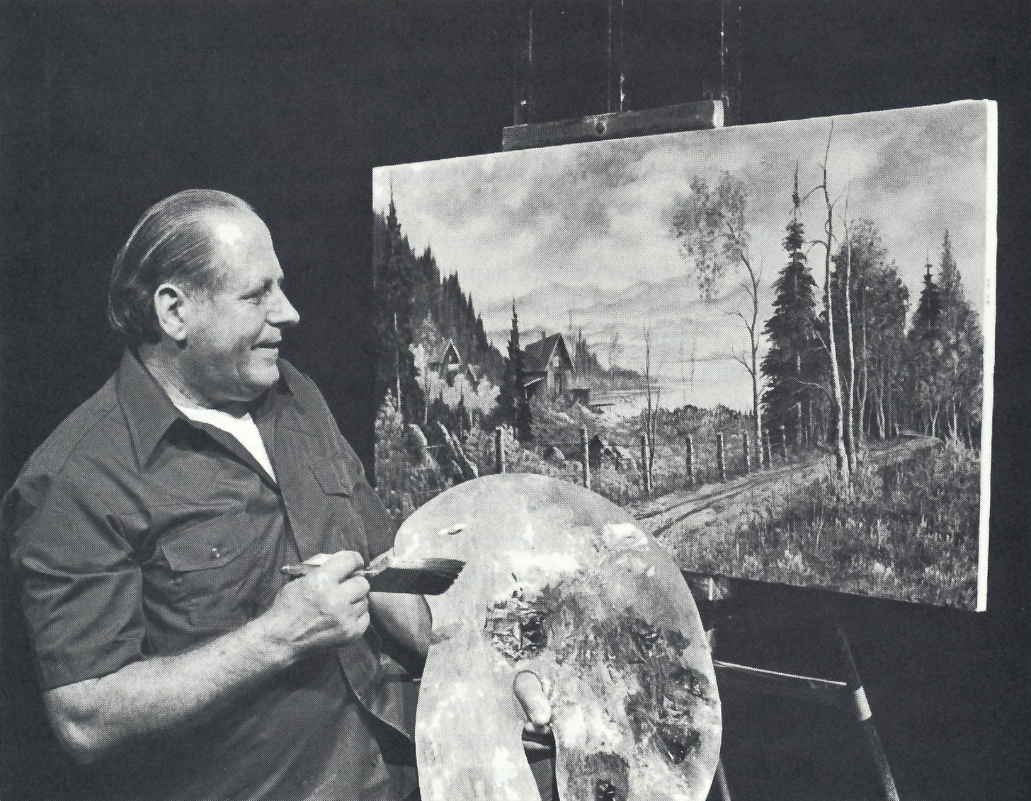 Bill Alexander is smiling and standing in front of a landscape painting with a palette in his hand