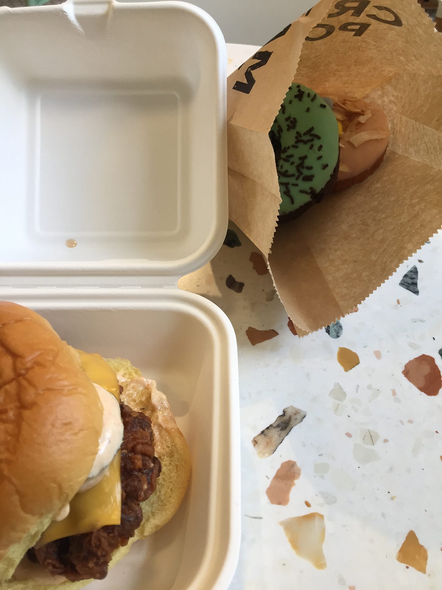 From above, a fried chicken sandwich with cheese, spicy mayo, and pickles in a white paper takeout box. Next to it in a brown paper bag with "Cream Pony" in block letters are two donuts, chocolate with green icing and chocolate sprinkles, and vanilla with pink icing, toasted coconut, and mini eggs in the centre.
