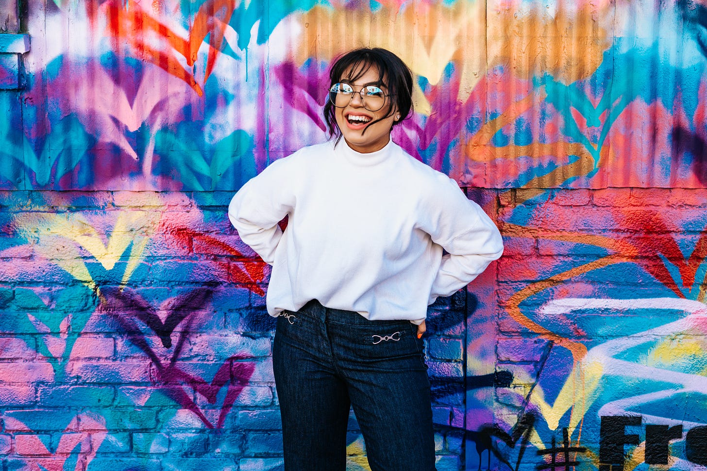 woman wearing a white sweatshirt standing in front of a colorful wall. She is smiling and wearing glasses.