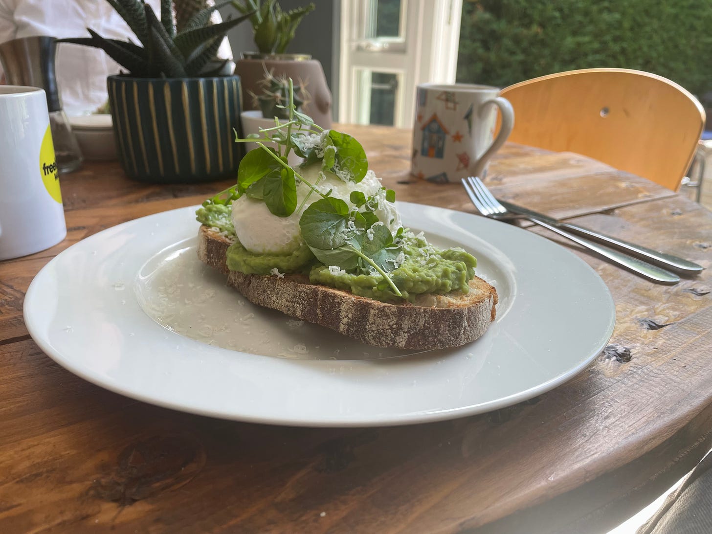 Piece of crusty toast holding broad bean puree, poached egg and a few cress leaves. Toast is served on a white round plate on a wooden table. A mug and cutlery are placed either side of the plate. 