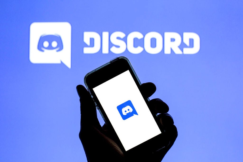 Photo illustration of a hand holding up a phone showing the Discord logo against a backdrop of another Discord logo. (Thiago Prudêncio / Getty Images)