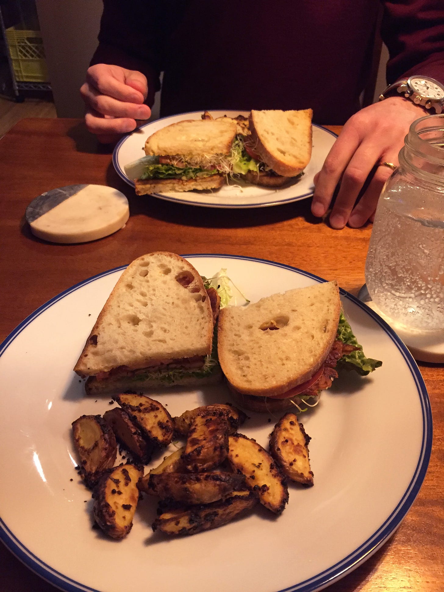 Two halves of a tempeh BLT with sprouts sit on a plate next to a small pile of mustard-roasted potatoes. The same food is on a plate at the other side of the table, with Jeff's hand on the table beside the plate.
