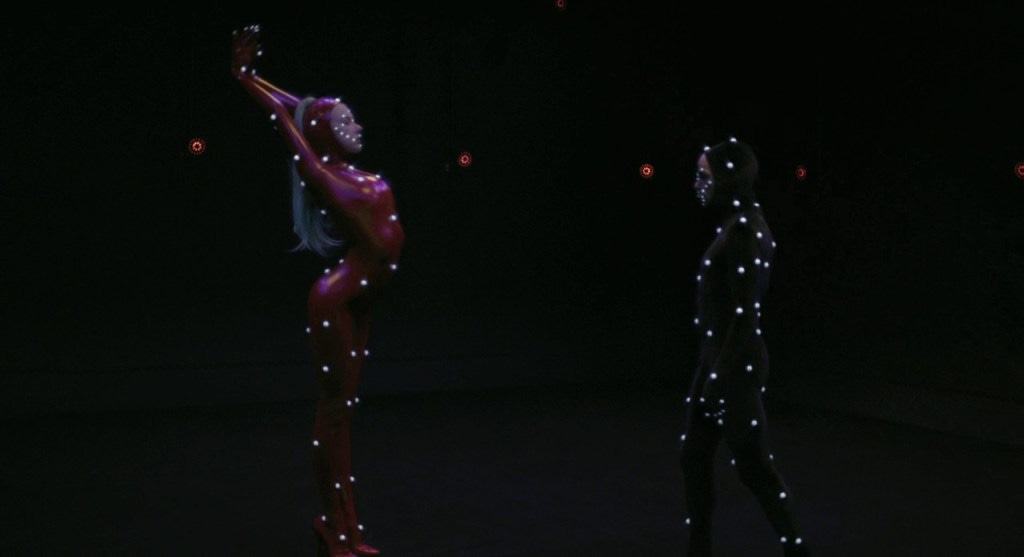 A film still from Holy Motors showing two people in mocap suits performing in a dark room. 