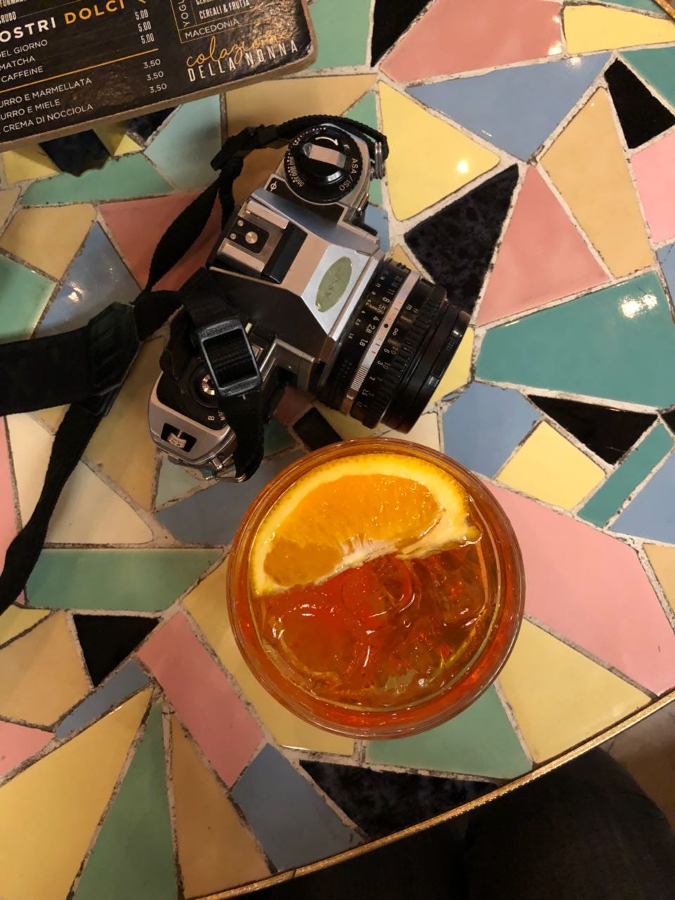 Eine analoge Kamera und ein Glas Aperol Spritz mit einer Orangenscheibe darin sind von oben fotografiert, die Tischplatte besteht aus einem bunten Mosaik.