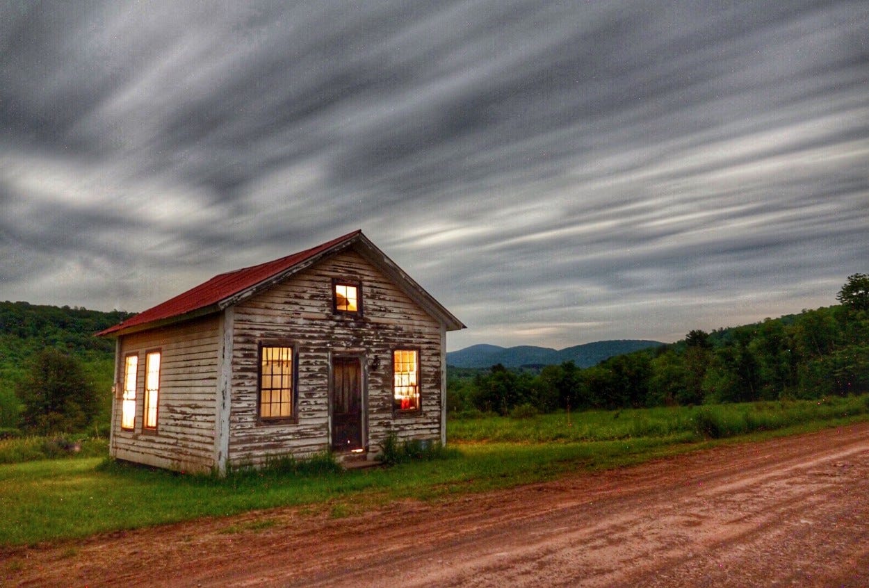 Exterior of an old one room schoolhouse at twight light. Red dirt road in front under a sky of streaking clouds.