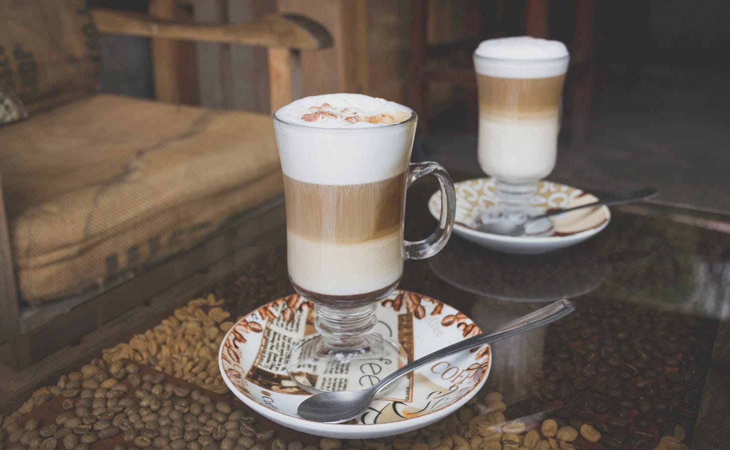 Two tall lattes in clear glasses on a glass coffee table. Coffee beans of various roast colors are underneath the glass and a chair with a coffee burlap sack converted to a chair cushion is in the background.