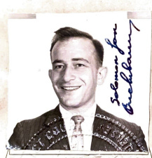 A passport-size photo of a young man, about 27, smiling broadly. He wears a dark suit jacket and bright shirt and tie. He has dark hair and broad features. The photo is signed "Solomon Jan Buchsbaum"