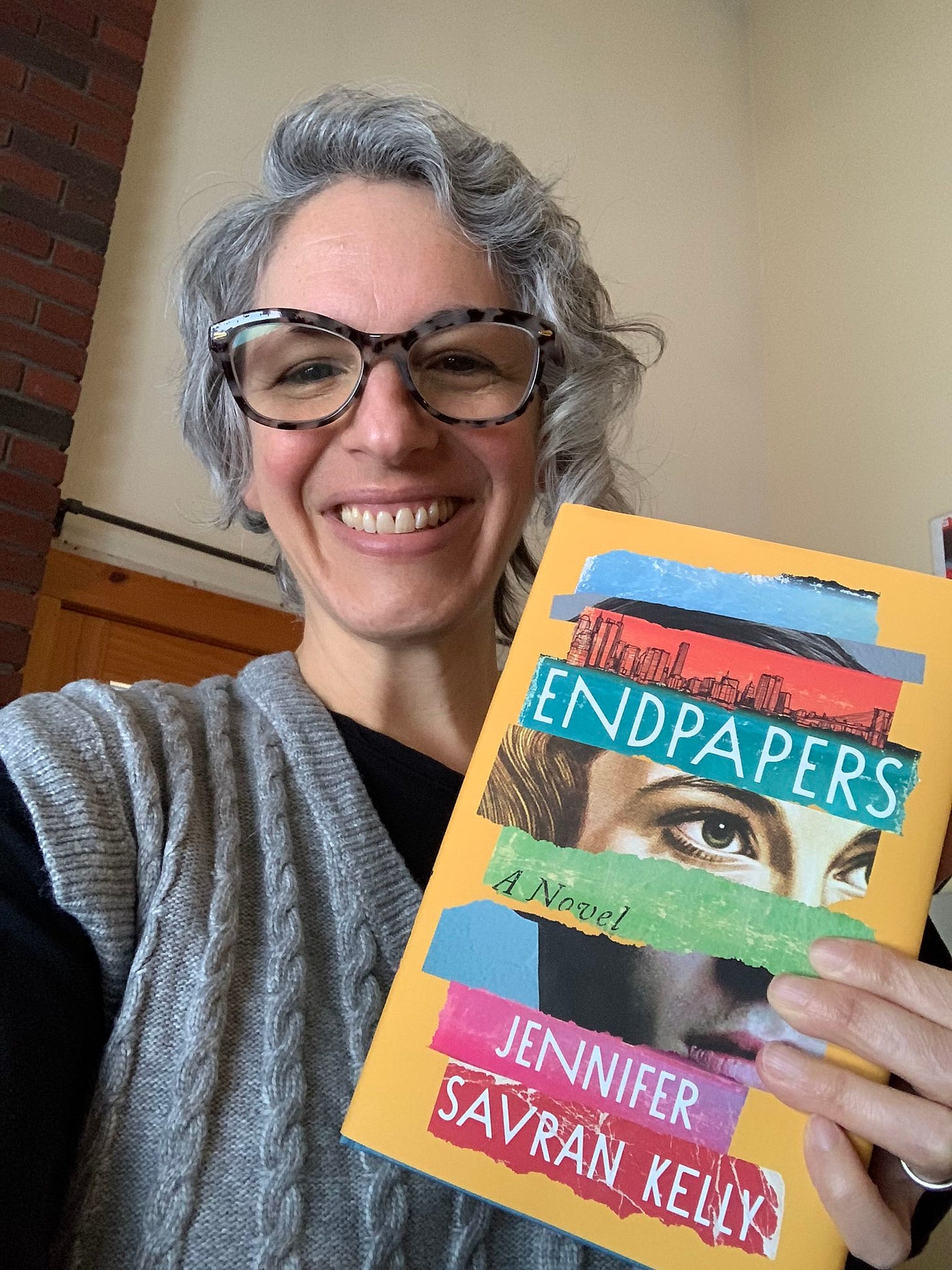 Selfie of me holding a copy of ENDPAPERS: A NOVEL by Jennifer Savran Kelly. I have short, curly gray hair and glasses.