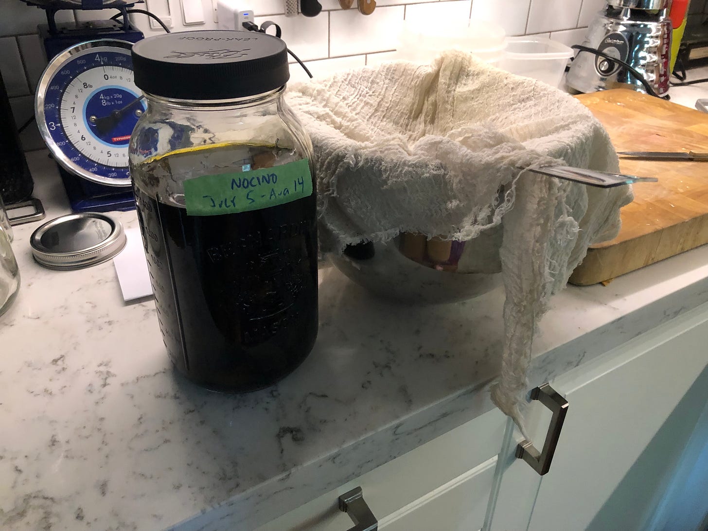 A large jar of nocino, about to be poured through a strainer lined with cheesecloth into a large bowl