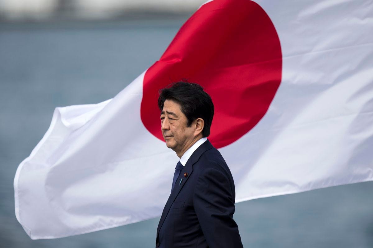 Shinzo Abe stands in a dark suit, in profile, against a massive Japanese flag furrowed by wind behind him. 