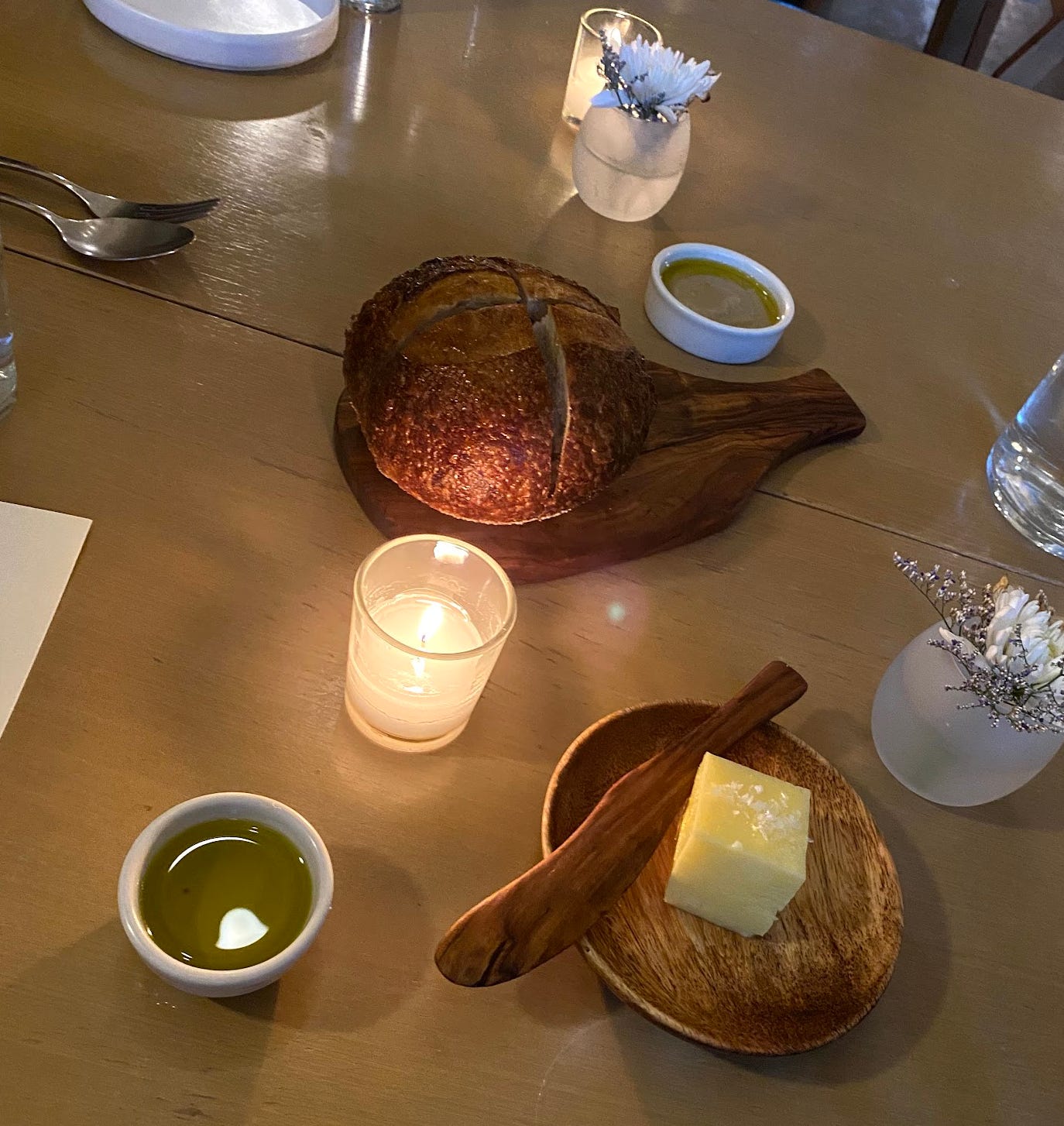 a round brown loaf, cut roughly into quarters, sits on a wooden serving board. next to it is a cube of golden butter, flecked with salt. the table is lit by two tiny candles, reflected in a small dish of olive oil. two tiny cups hold one white chrysanthemum (i think) each. 
