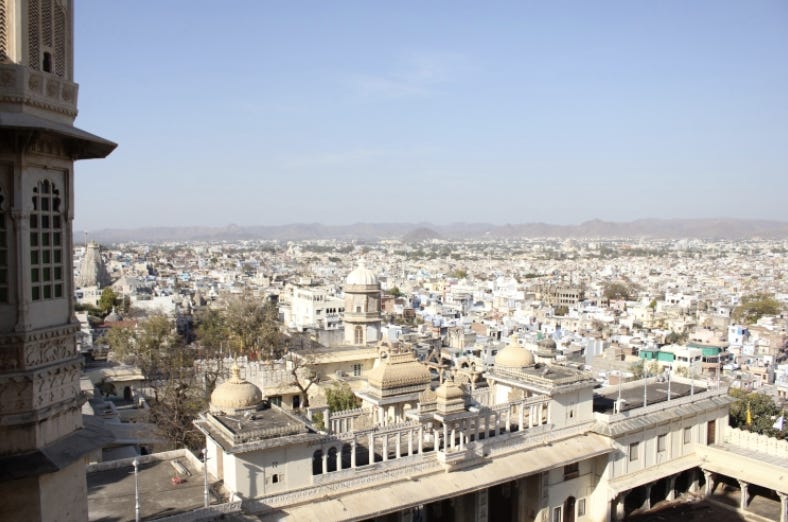 A city during the day filled with bright white buildings. Some of the buildings have domes. In the background there are mountain plains. Above is a light blue sky.