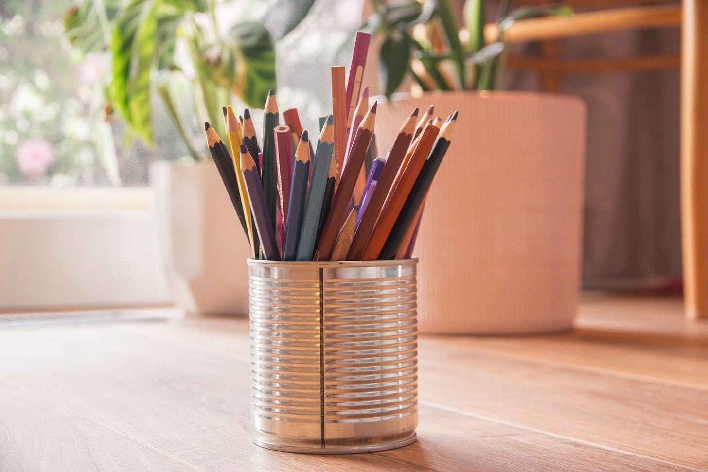 Pot of coloured pencils on a wooden desk by a window in daytime.