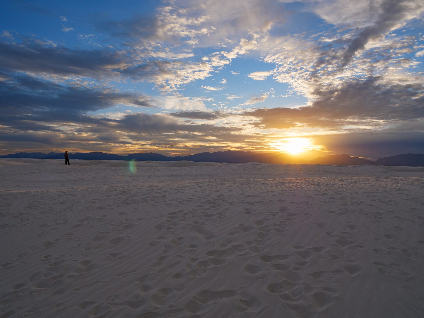 A wide view of White Sands National Park. White powdery sand fills the lower half of the photo. In the sand you can see contours and striations created by the winds and the footsteps of visitors. On the left, near the edge of the visible sand, stands a solitary figure, a park ranger. They are looking towards the distant mountains in the background. They are purple and jagged. Just above them, a bright, golden sun is setting in a blue sky interspersed with clouds at different altitudes. The low clouds are deep grey, their edges tinged with rays of the sun. Higher up, the clouds are white.  A sliver of the sand is touched by the sunlight and is colored golden. A small green lens flare (an optical distortion caused by pointing the camera at the sun) floats near the park ranger.