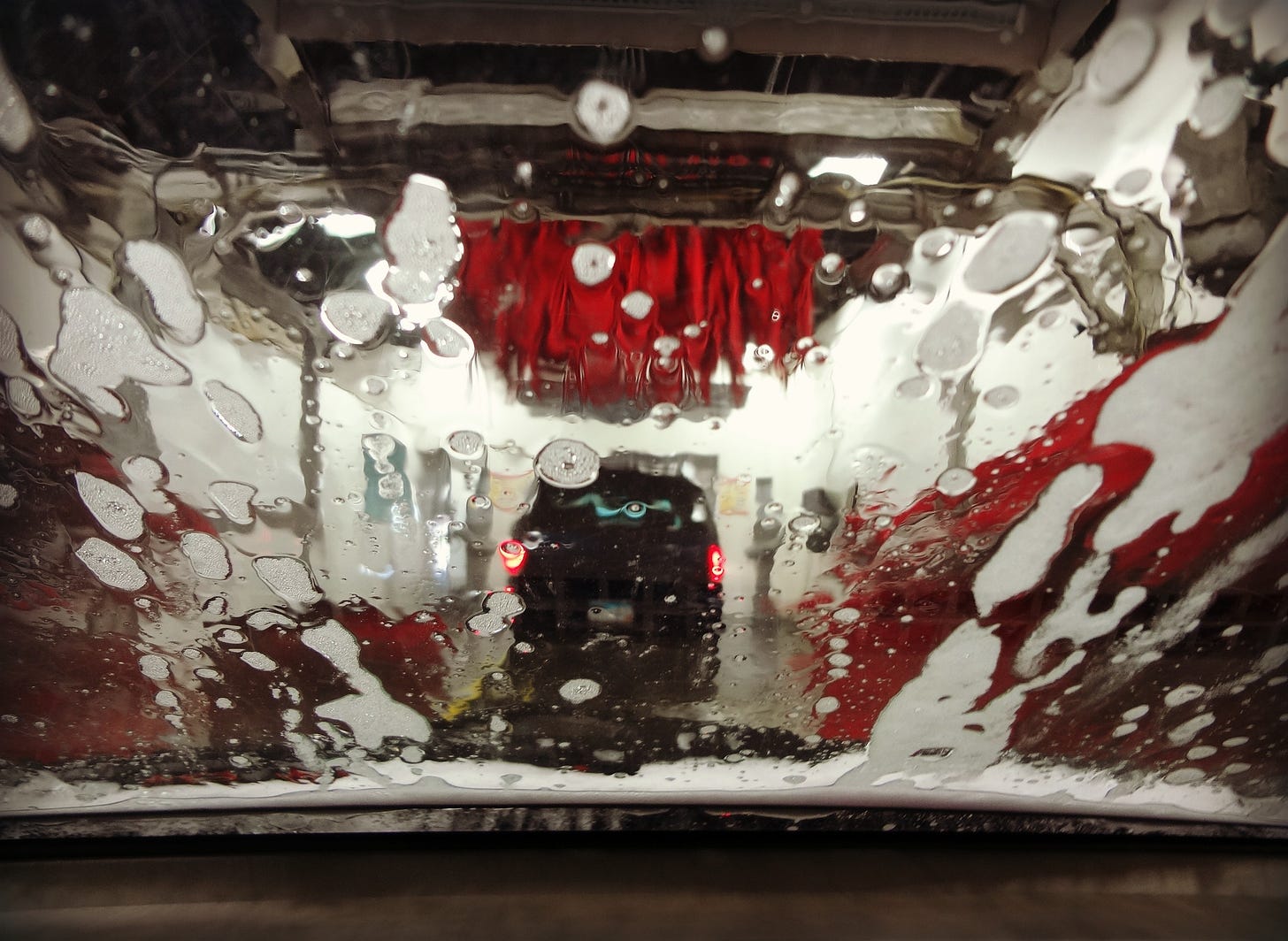 Sudsy water dripping down the windshield of a car in an automated car wash with red bristled brushes