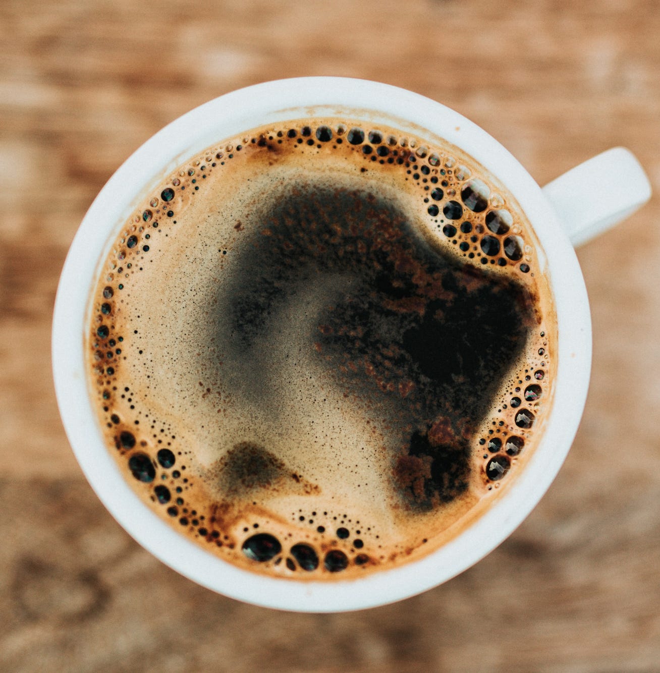 On overhead view of a black coffee in a white ceramic coffee mug resting on a blurred wood countertop.