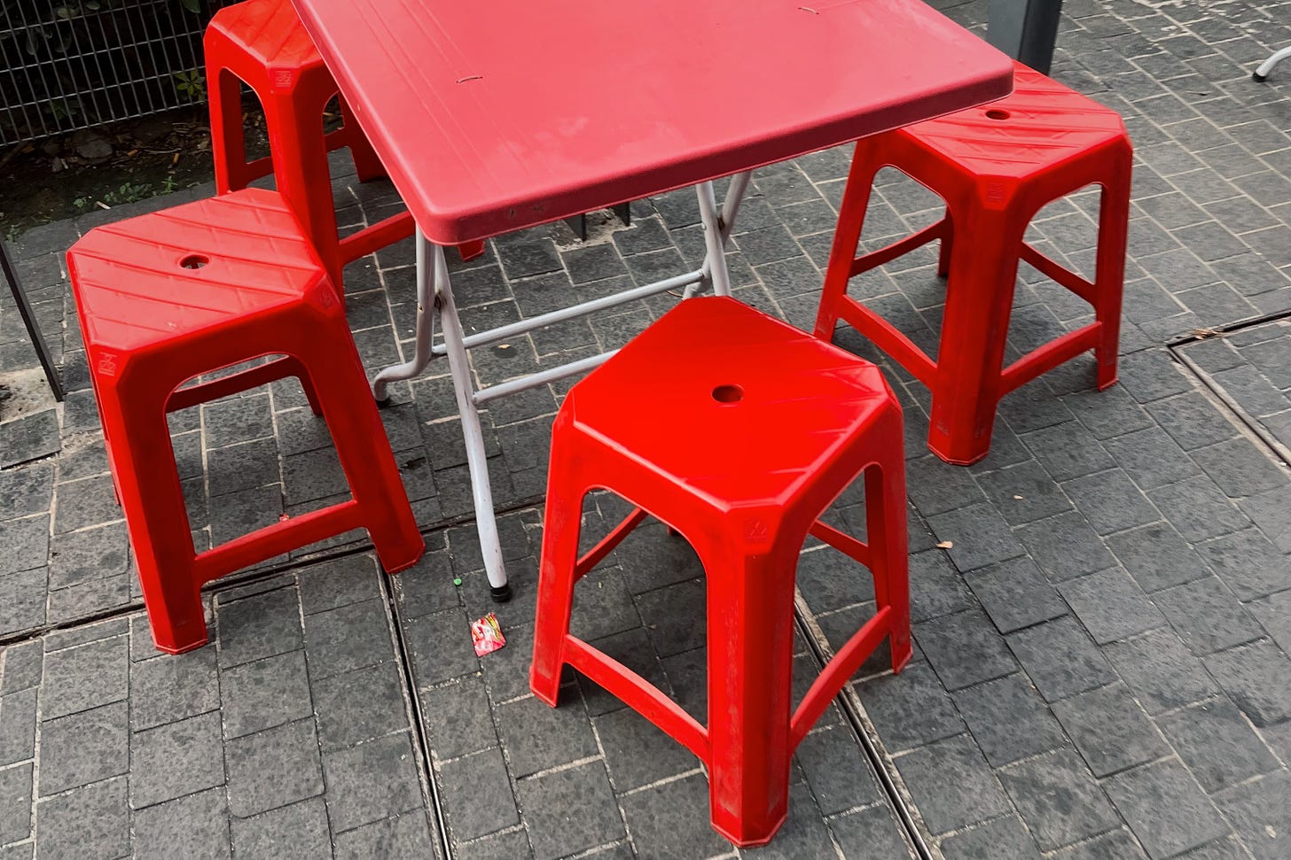 a red fold-out table on a grey-paved street, surrounded by four red plastic stools. the photograph is taken in passing, at a high angle, with no other background details apparent