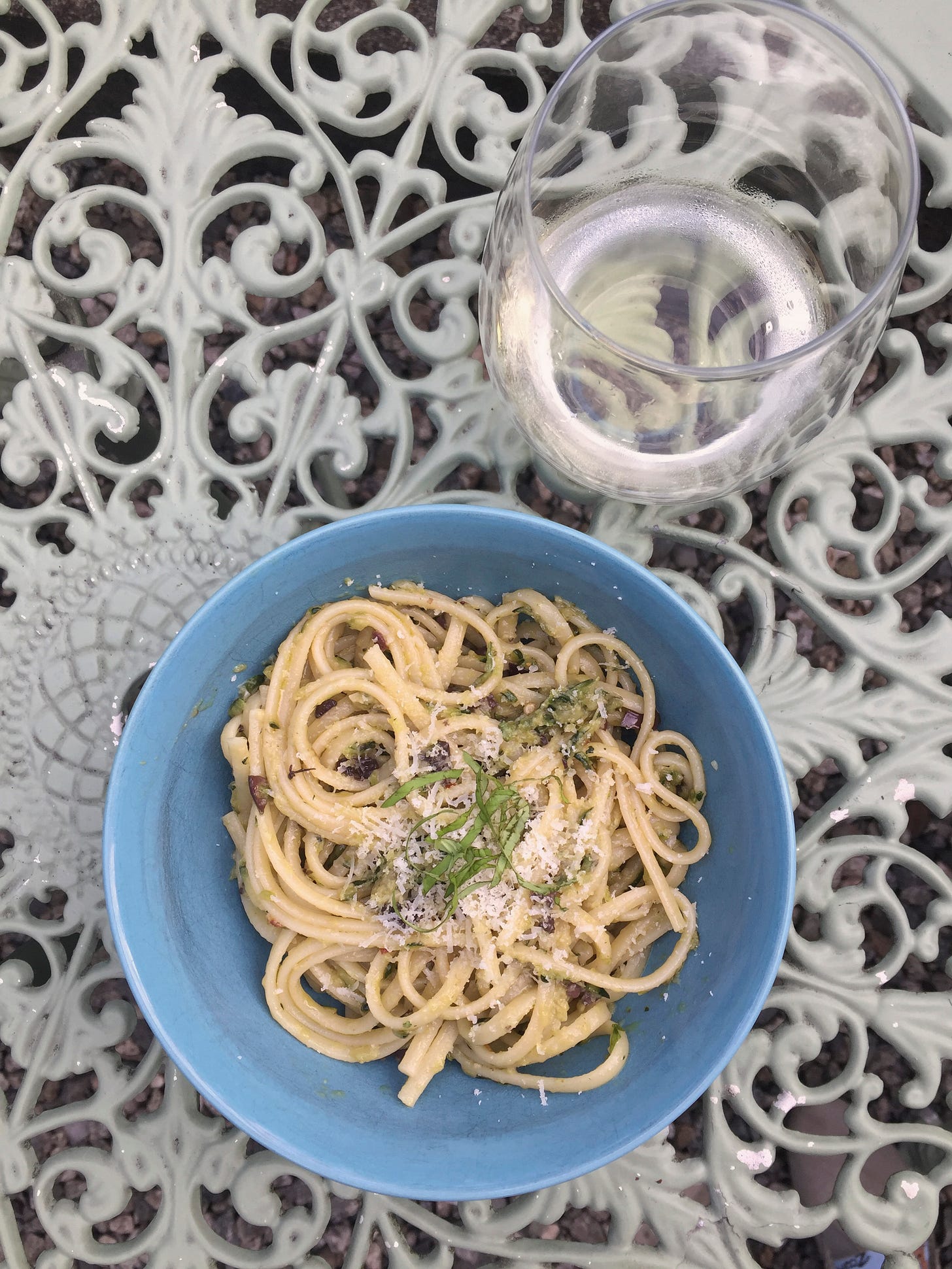 On an outdoor table, a stemless glass of white wine sits at the upper right corner of a blue bowl filled with linguine, and topped with parmesan and basil.