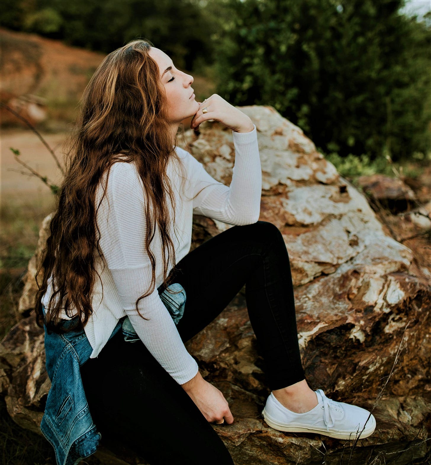 girl with long brown hair wearing long sleeve white top sitting on boulder with hand under chin and eyes closed