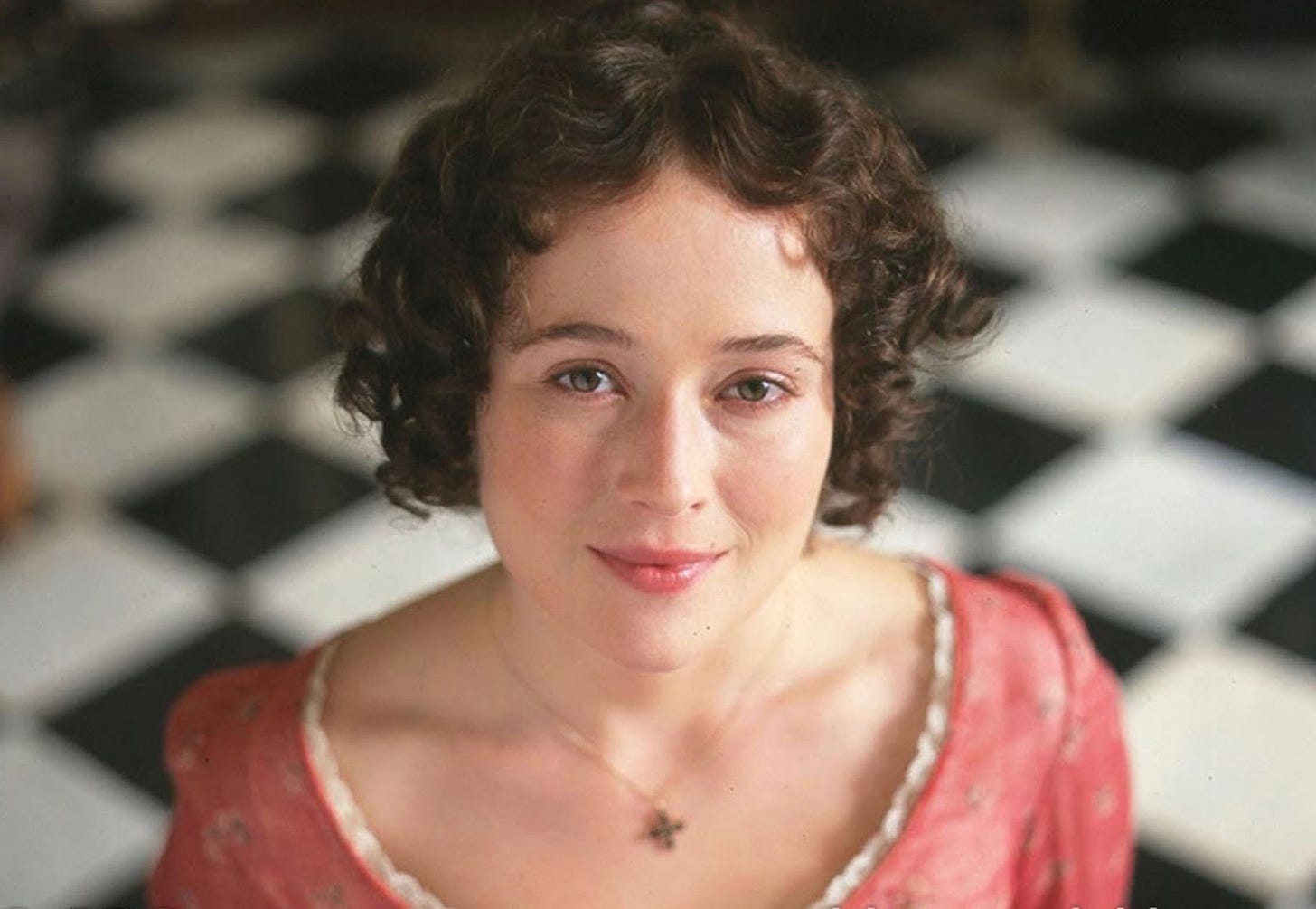 The actor Jennifer Ehle, portraying Elizabeth Bennet, gazes straight up to the camera. She wears a mauve Regency gown with white lace trim and a necklace, and a bemused but serene expression. In the background is a black and white checked floor pattern. 
