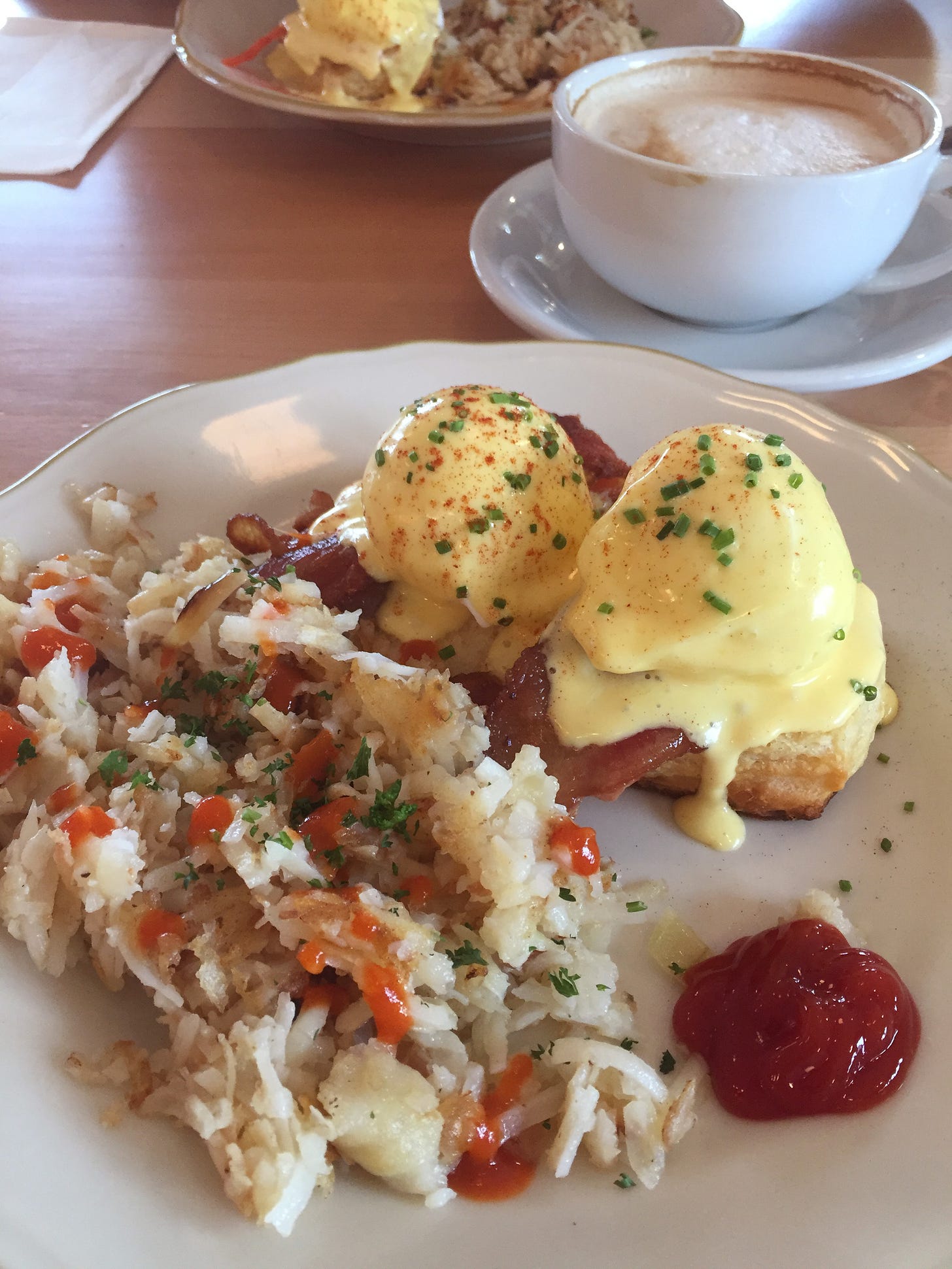On a white plate with gently scalloped edges is a pile of shredded hash browns dotted with hot sauce, next to a small puddle of ketchup and in front of two eggs benedict, dusted with paprika and chives. A large cup with most of a latte rests on a sauce behind it, and a second plate of food is just visible in the background.