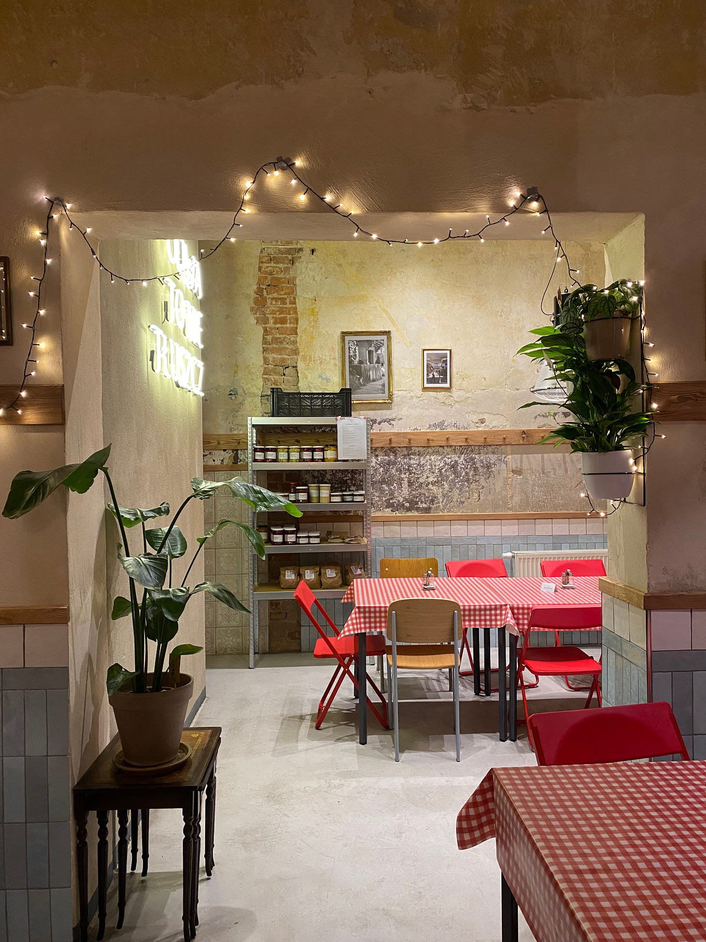 Another view of the restaurant from before. There is a bare brick wall with occasional plaster. Small tables sit in the background with the checkered tablecloths and folding chairs. In the foreground, there are fairy lights and house plants.