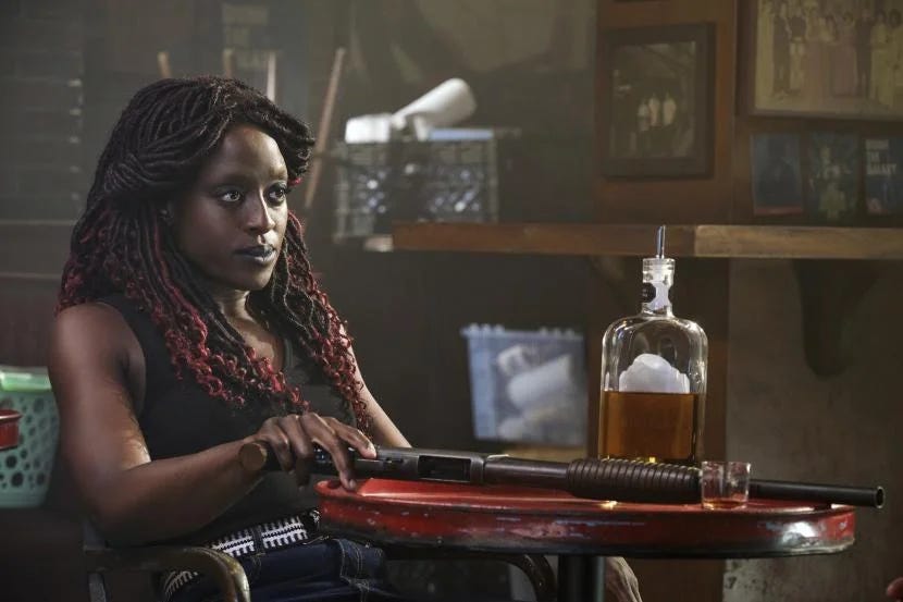 This image shows Young Guinan (played by Ito Aghayere) sitting at a table inside her 10 Forward Avenue bar with her right hand grasping the butt of a shotgun that lies on that table. A bottle of whiskey sits next to the shotgun. This image shows Young Guinan (played by Ito Aghayere) sitting at a table inside her 10 Forward Avenue bar with her right hand grasping the butt of a shotgun that lies on that table. A bottle of whiskey sits next to the shotgun.