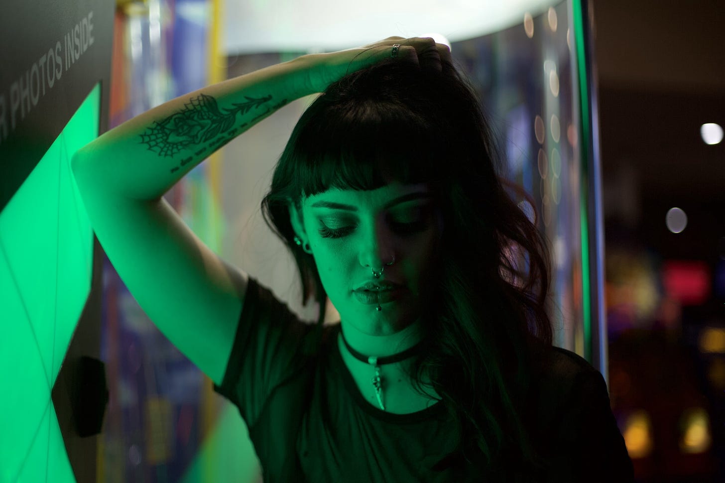 A young woman with dark hair and tattoos is standing in an arcade and painted green with light.