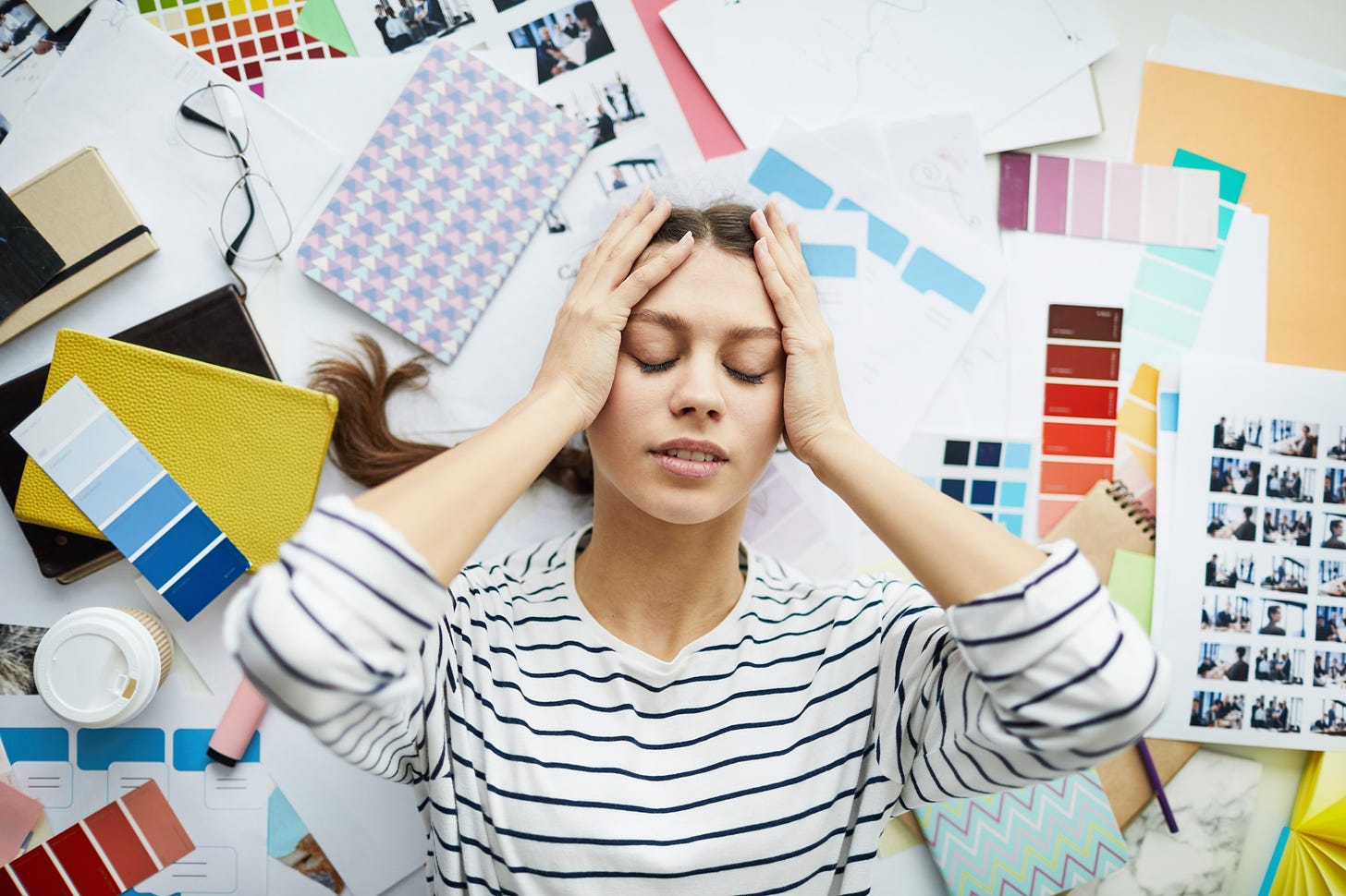 A stressed young woman lays in a pile of clutter with her head in her hands and her eyes closed.