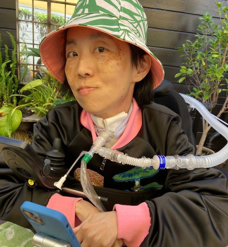 I’m at Stable Cafe, in San Francisco, where they have a gorgeous outdoor patio with lots of greenery. I am wearing a sunhat with a leaf print and a black jacket with pink trim. My phone is in front of me on a holder so I can communicate.