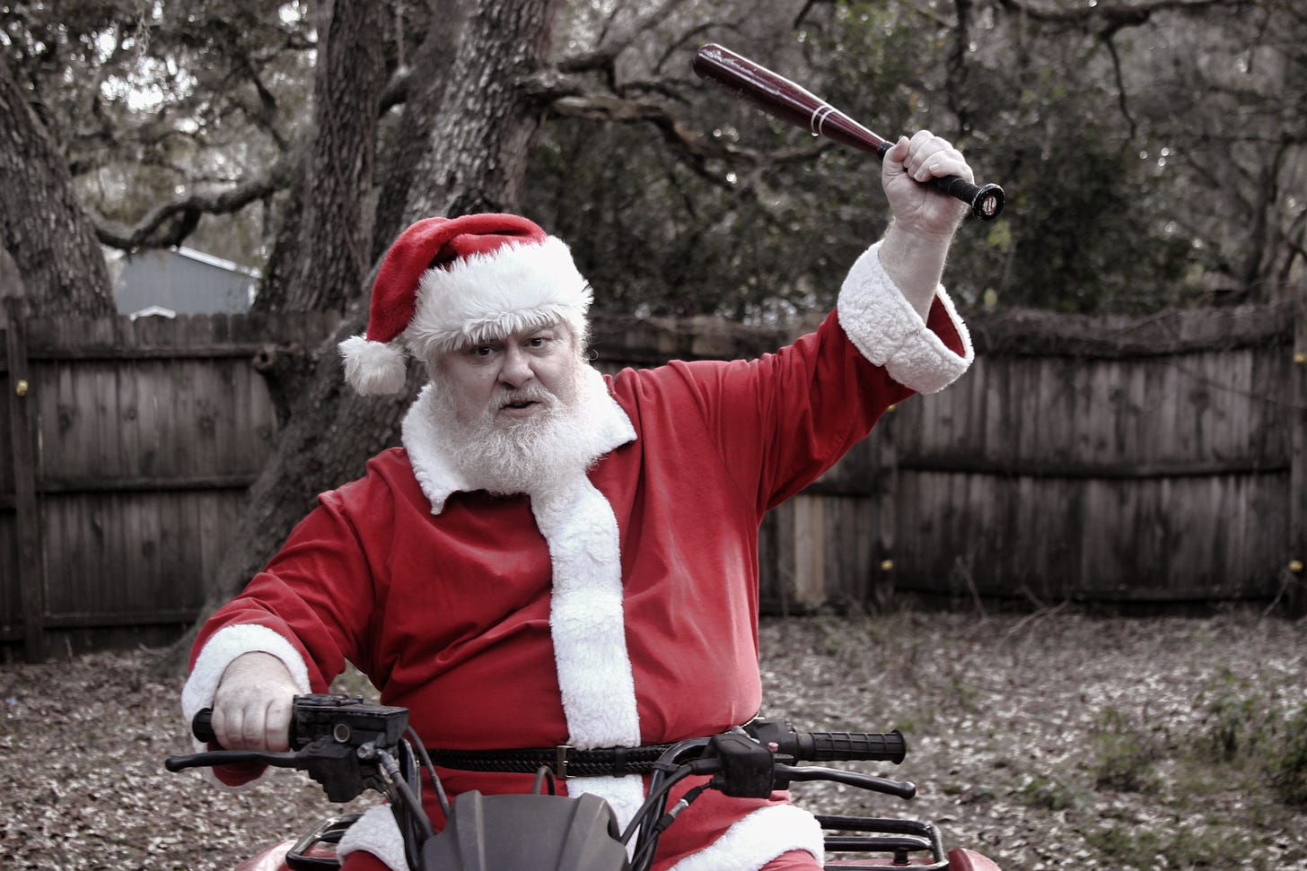 An angry looking santa is on a bicycle and he is raising a bat in his hands at whoever is taking the picture. In the background are bare trees and dead leaves and a garden fence.