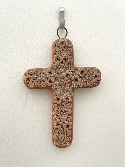 Ceramic cross with decorative plants carved into it hanging on a white wall.